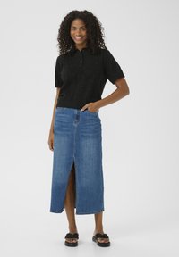 Schwarzes gestricktes Polohemd mit Knöpfen und kurzen Ärmeln, kombiniert mit einem hoch taillierten Maxispiel aus blauen Jeans mit einem vorderen Schlitz. Schwarze Sandalen.