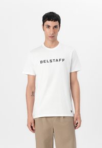 Fehér pamut póló, amelyen a "BELSTAFF" logó vastag fekete betűkkel szerepel. Szabványos fit, rövid ujjak és kerek nyakú.