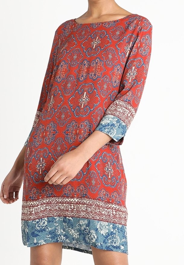 Femme portant une robe longueur genou avec motifs floraux rouges, manches trois-quarts, et bordures décoratives bleues et blanches aux manches et à l'ourlet.