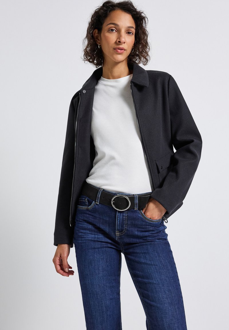 Nainen, jolla on kihara hius, pukeutuneena mustaan vetoketjulliseen takkiin, valkoiseen paitaan, tummansinisiin farkkuihin ja mustaan vyöhön, jossa on pyöreä hope buckle.