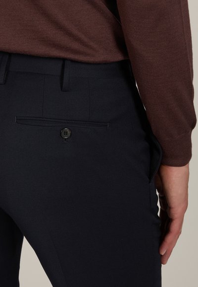 Close-up of a person wearing dark navy trousers with a buttoned back pocket and a brown long-sleeve top, hand resting by their side.