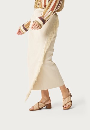 Mujer vistiendo una falda midi color crema con detalle de flecos, camiseta de manga larga a rayas y sandalias beige de tacón ancho y punta abierta.