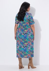 Kvinde med langt mørkt hår iført en flerfarvet blomsterprint midi-kjole og lysebrune ruskindsstøvler med hæl, stående mod en hvid væg med ryggen til.