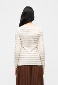 Γυναίκα με μακριά σκούρα μαλλιά που φοράει λευκό τοπ με μακριά μανίκια και οριζόντιες μπεζ ρίγες και καφέ πτυχωτή φούστα, με την πλάτη στραμμένη, σε λευκό φόντο.