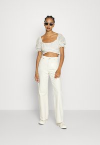 Top branco cropped com mangas bufantes, padrão de ilhós, combinado com jeans brancos de cintura alta e ténis brancos. Acessórios incluem óculos de sol.