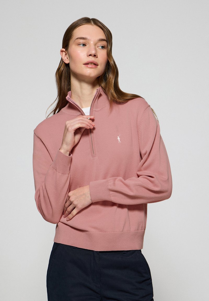 Pull light rose avec un col zippé semi-ouvert, des poignets et un ourlet côtelés. Présente un petit logo brodé sur la poitrine. Texture lisse.