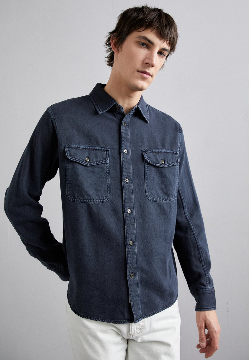 Mørkeblå denimskjorte med lange ermer, to brystlommer, knappestolpe og stikninger. Myk tekstur og uformell design.