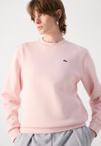 Ljusrosa sweatshirt med rund halsringning, långa ärmar och textilerad material. Har en liten grön logotyp på vänster bröst.