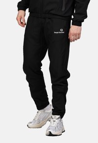 Schwarze Jogginghose aus leichtem Material mit elastischem Bund, seitlichen Taschen und einem weißen Logo am Unterschenkel. Kombiniert mit weißen Sneakers.