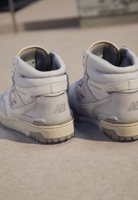 Balti aukštakulniai New Balance sportbačiai, padėti šalia vienas kito ant lygaus lauko betono paviršiaus dienos šviesoje.