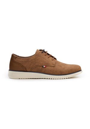 Zapatos de ante marrón con cordones y suela de goma blanca. Incluyen un puntero redondeado y un pequeño logo en rojo, blanco y azul en el costado.