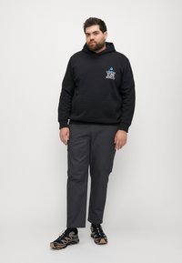 Schwarzer Hoodie mit grafischem Text auf der Brust, kombiniert mit dunkelgrauen Cargo-Hosen und schwarzen Sportschuhen mit beigen Akzenten.