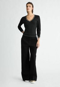 Zwarte gebreide top met een V-hals en verticale strepen, gecombineerd met een wijde zwarte broek. Witte en gouden sneakers maken de outfit compleet.