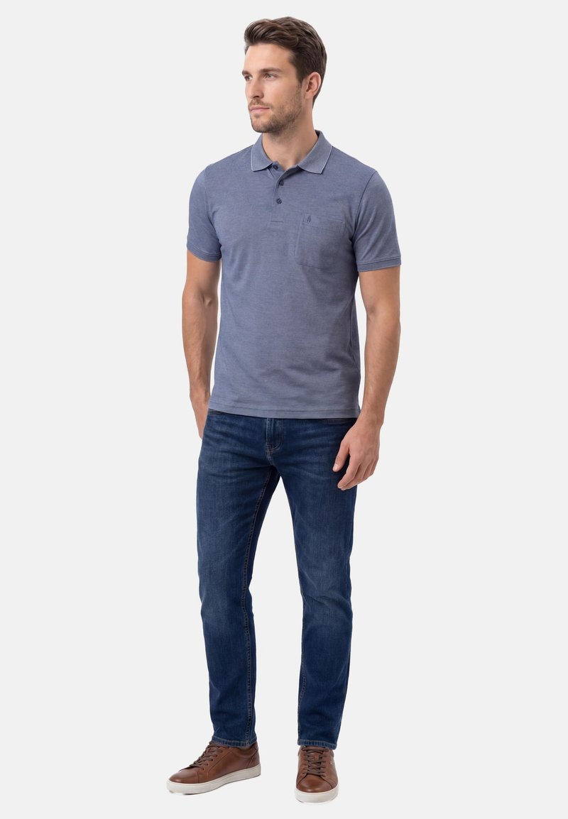 Homme debout, portant un polo à manches courtes bleu, un jean bleu foncé et des baskets marron, faisant face légèrement vers la gauche.