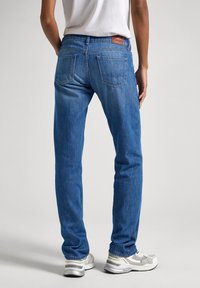 Jeans de mezclilla azul claro con un corte ajustado, dos bolsillos traseros y un parche de cuero marrón en la cintura. Llevados con zapatillas blancas y grises.