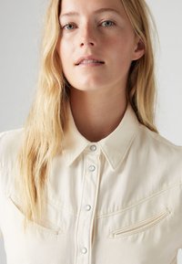 Camicia in denim color crema con colletto a punta, chiusura con bottoni e due tasche frontali con patte curve. Dettagli sottili di trama e cuciture.