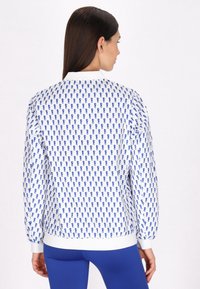 Hanorac alb cu un model albastru repetitiv, manșete și tiv ribbed, și o croială clasică. Nu sunt vizibile detalii pe față.