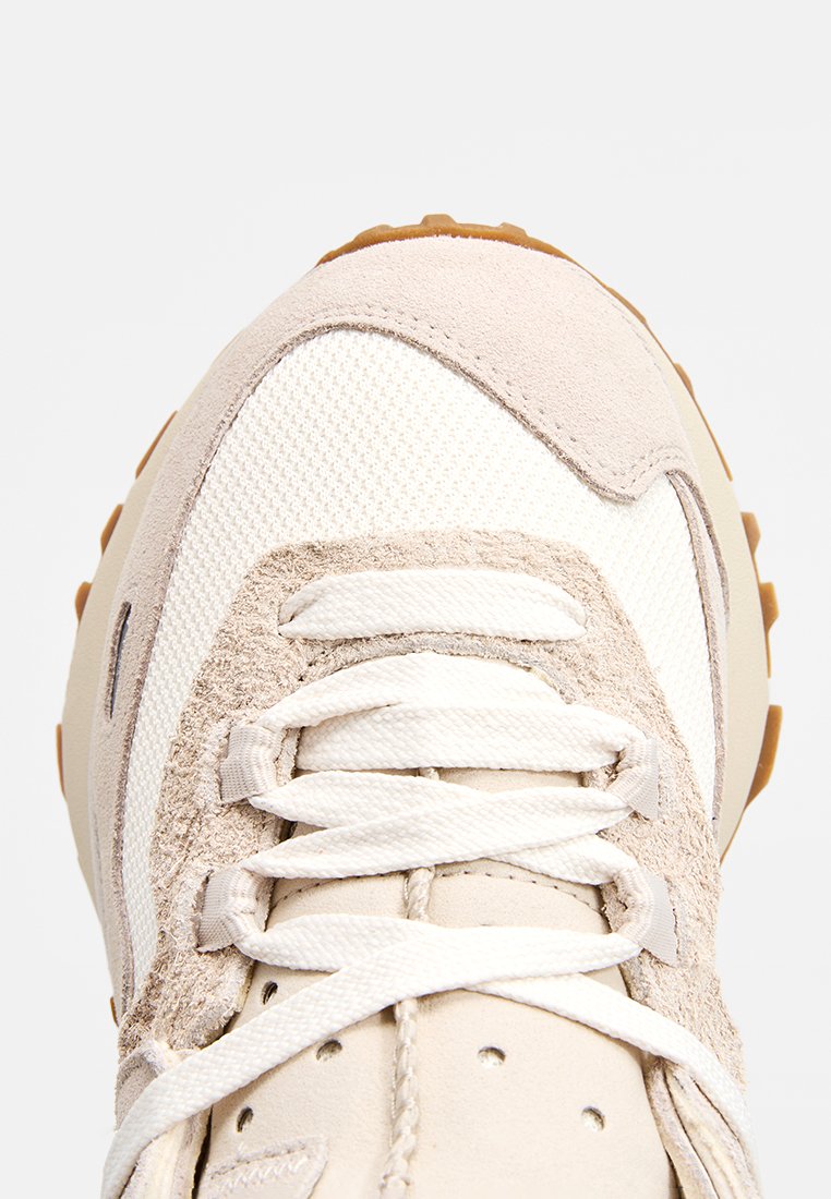 Sneaker beige clair présentant un mélange de daim texturé et de matériaux en maille, bout rond, lacets plats et semelle en caoutchouc avec rainures de traction.