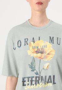 Gaiši zaļa lielizmēra T-krekls ar dzeltenu ziedu grafiku, kurā ir teksts "SOW LOVE" un "ETERNAL Sunshine" tumšā burtu stilā.