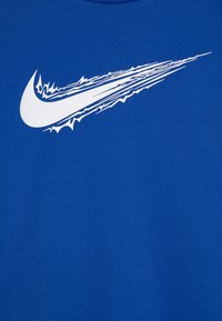 Kék Nike póló, amelyen egy feltűnő fehér swoosh logó látható, dinamikus kialakítással, sima, texturált anyag háttérben.