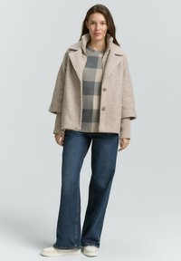 Beige, gestructureerde buitenjas met grote revers, twee knopen en een gelaagd grijs en crème geruit sweater eronder. Donkere jeans en lichte schoenen.