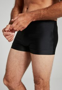 Schwarze Schwimmshorts mit einer glatten, glänzenden Textur, die eine tiefe Passform und ein minimalistisches Design aufweisen. Das Model passt den Bund an.