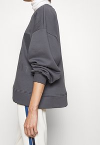 Sudadera gris de gran tamaño con puños y dobladillo acanalados, que presenta un diseño texturizado en la parte frontal. Combinada con pantalones crema y rayas azules.