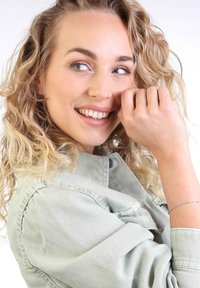 Mujer con cabello rizado en una chaqueta de mezclilla verde clara sonríe, mostrando una delicada pulsera de plata en su muñeca, contra un fondo blanco liso.