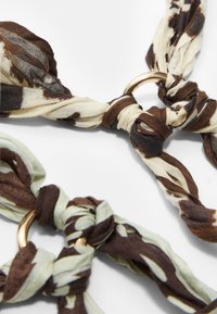 Tessuto marrone e crema con motivo a disegni annodati e un anello in metallo dorato, caratterizzato da un materiale morbido e leggero e texture variabili.
