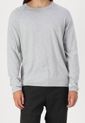 Person trägt ein schlichtes hellgraues Langarmshirt und eine dunkle Hose, dargestellt vom Hals bis zu den oberen Oberschenkeln vor einfarbigem Hintergrund.