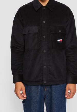 Schwarze Cordjacke mit Knopfleiste und zwei Brusttaschen, eine mit kleinem rot-weiß-blauem Tommy Jeans Logo-Patch, getragen zu blauen Jeans.