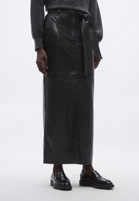 Mustast nahast midi seelik siledal tekstuuril, vöö detailiga ja sirge lõikega, kombineeritud läikivate mustade loafersidega neutraalsel taustal.