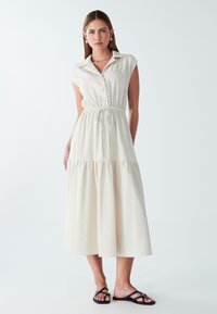 Nupuk, ilma varrukatega, kreemivärvi kleit elastse vöökoha ja kihilise seelikuosa ning sujuva kangaga, millel on krae ja kogumisseatud detailid.