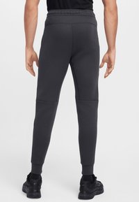 Pelēki sporta bikses ar konisku griezumu, sānu paneļiem un elastīgu jostu. Valkātas kopā ar melniem sporta apaviem.