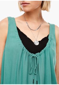Top sin mangas en color turquesa con cuello ajustable, superpuesto a un top negro. Presenta una tela suave y fluida con un corte relajado. Incluye un collar de plata.