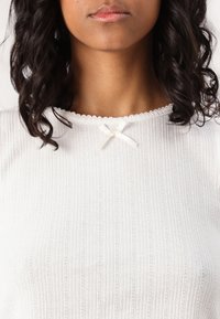 Top bianco a maglia con texture a coste orizzontali, scollatura a scallop e un piccolo dettaglio in stoffa a forma di fiocco al centro anteriore.