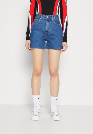 Kvinde iført blå denimshorts, en sort langærmet top med røde og hvide striber, hvide sokker og hvide sneakers med røde og blå accenter.