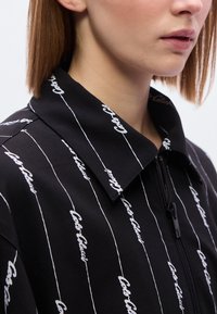 Μαύρο μπουφάν με φερμουάρ και γιακά, με λευκές διαγώνιες γραμμές και επαναλαμβανόμενα μο τίβα κειμένου. Απαλή υφή, casual σχέδιο.