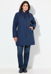 Parka impermeabile blu navy con cappuccio, dotata di coulisse rossa, bottoni a pressione e due tasche laterali. Indossata sopra un paio di jeans blu e scarpe nere.