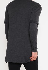 Mann, der ein dunkelgraues, langärmeliges Schichthemd trägt, von hinten gezeigt, hält den Saum des äußeren Hemdes mit einer Hand.