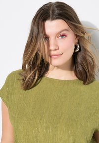 Blusa verde texturizada con mangas cortas, que presenta un brillo sutil. Acentuada con pendientes de aro plateados y cabello ligeramente peinado.