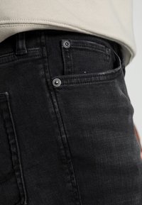 Vaqueros de denim negro con textura suave, presentando un corte ajustado, bolsillos delanteros y detalles en herrajes metálicos. Detalle de costuras visibles.