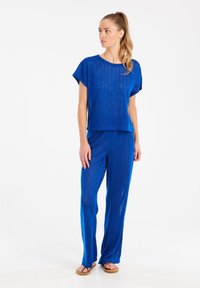 Mėlynas megztas dviejų dalių komplektas, kuriame yra trumpomis rankovėmis viršus ir laisvi kelnės su deimanto raštais; derinamas su smėlio spalvos sandalais.