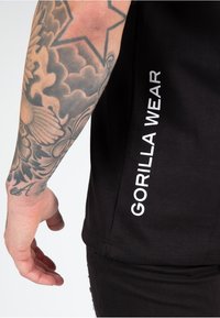 Juodas marškinėliai su balta "GORILLA WEAR" užrašu šone. Artimas vaizdas, rodantis ranką ir tatuiruotą ranką. Minkšta medvilnė, trumpomis rankovėmis.