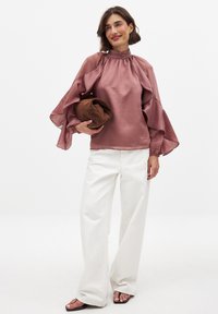 Bordeaux satijnen blouse met geflarde, doorschijnende mouwen en een hoge halslijn, gecombineerd met witte broek met wijde pijpen en een bruine clutch.