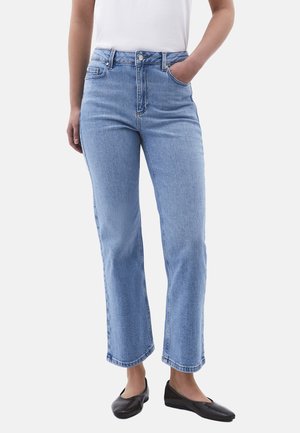 Mujer con jeans de pierna recta color azul claro y una blusa blanca, con una mano en el bolsillo y zapatos planos negros, sobre un fondo liso.