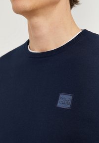 Ung person som bär en marinblå tröja med rund hals och en liten "Polo Club"-patch på vänster bröst.