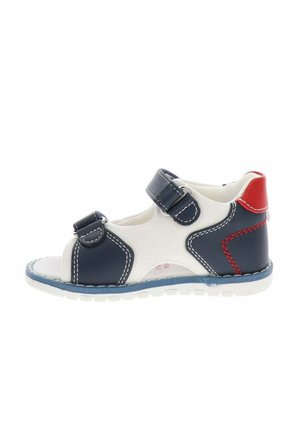 Sandalia infantil con punta abierta con tiras azul marino, base de tela blanca, detalle rojo en el talón y cierres de velcro ajustables sobre una suela blanca.