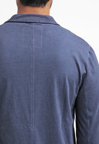 Uomo con indosso una camicia di cotone blu visto da dietro, con una cucitura centrale e un dettaglio rettangolare cucito sotto il colletto.