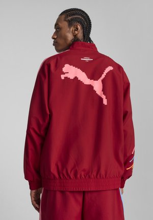 Red windbreaker jacket featuring a large pixelated pink logo on the back, elastic hem, and colorful stripes on the sleeves.